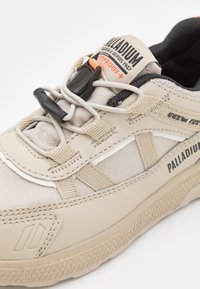 Adidași bej cu un upper din material texturat, talpă din cauciuc, șireturi elastice și un capac de tras negru; prezintă un logo imprimat și un design impermeabil.