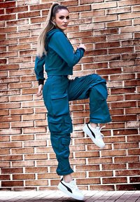 Teal cropped hoodie en bijpassende cargobroek met grote zakken, gecombineerd met witte sneakers. Glad textuur, atletische pasvorm, tegen een bakstenen muur.