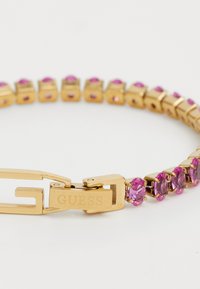Kuldtooni käevõru, millel on roosad rhinestone'id ovaalsetes asetustes, ja lukuga, millel on "GUESS" logo ning painutatav, paindlik disain.
