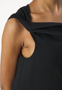 Gros plan d'une personne portant un haut noir sans manches avec un détail de bretelle torsadée sur l'épaule.