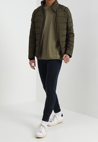 Olivgrön puffjacka med dragkedja fram, matchad med en mörkblå skjorta och joggers. Skorna är vita sneakers med beige detaljer.