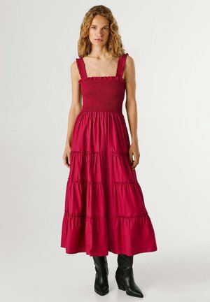 Vrouw draagt een mouwloze, gelaagde rode midi-jurk met een gerimpelde halslijn en zwarte, puntige mid-kuit laarzen, staand tegen een effen achtergrond.