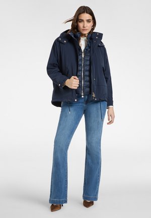 Frau trägt mehrere navyblaue Jacken, hellblaue Bootcut-Jeans und braune spitze Schuhe, steht vor weißem Hintergrund.