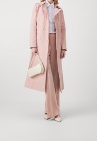 Pitkä, vaaleanpunainen trenssitakki, jossa kaksiriviset napit, puettu valkoisen paidan ja vastaavien vaaleanpunaisten housujen päälle. Kantaa beigeä käsilaukkua.