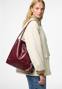 Giovane donna con lunghi capelli biondi che indossa una giacca beige e jeans blu, porta una grande borsa a tracolla in pelle lucida di colore rosso scuro.