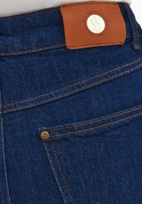 Detailní záběr na zadní kapsu tmavě modrých džín s oranžovým švem a hnědou koženou značkovou nášivkou.