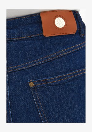 Zbliżenie na tylnią kieszeń ciemnoniebieskich dżinsów z pomarańczowymi szwami i brązową skórzaną metką marki.