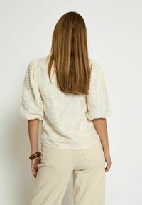 Cream-colored textured blouse with puffed sleeves, featuring a floral design. Worn with beige wide-leg pants.