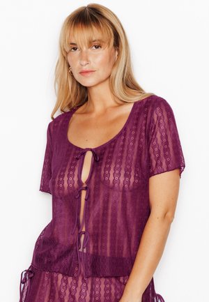 Donna con capelli biondi che indossa un top di pizzo viola trasparente a maniche corte, legato davanti con tre fiocchi, su sfondo bianco.