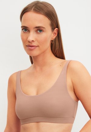 Young woman with straight light brown hair wears a beige sleeveless crop top and small gold hoop earrings against a plain background.