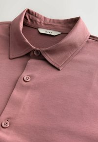 Mauve spalvos marškiniai su išsilenkusia apykakle, kuriuose matosi trys gumbai ir prekės ženklo etiketė vidinėje apykaklės pusėje.