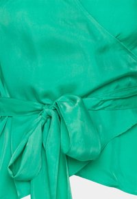 Haut cache-cœur en satin vert avec une taille nouée. Caractérisé par une texture lisse et un décolleté en V profond, avec une silhouette fluide et un ourlet asymétrique.