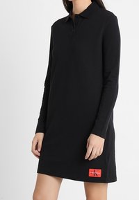 Žena ve černých dlouhých polo šatech s červeným logem Calvin Klein u lemu.