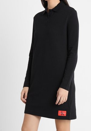 Kobieta ubrana w czarną sukienkę polo z długim rękawem, z czerwoną naszywką logo Calvin Klein blisko dołu.