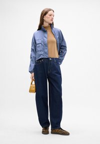 Chaqueta corta azul claro, jersey de cuello alto marrón claro, jeans sueltos azul oscuro y zapatillas de charol con estampado de leopardo. Sostiene un pequeño bolso amarillo. Fondo liso.