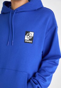 Kungsblå hoodie av mjukt tyg. Har en framficka och en svart fyrkantig logotyp med vita och blå detaljer. Snören i huvan.