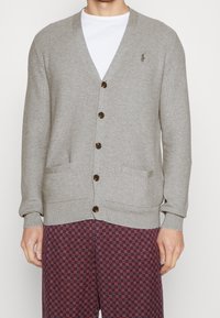 Ljusgrå cardigan med V-hals, träknappar, två framfickor och ett strukturerat mönster. Bärs över en vit skjorta och mönstrade byxor.