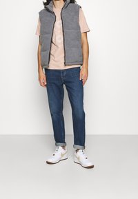 Gilet in pile grigio con collo alto, t-shirt rosa con grafica circolare, jeans blu e sneaker bianche con dettagli beige e logo verde.