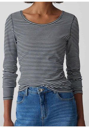 Femme portant un t-shirt à manches longues rayé noir et blanc horizontalement, rentré dans un jean bleu taille haute, debout devant un fond uni.