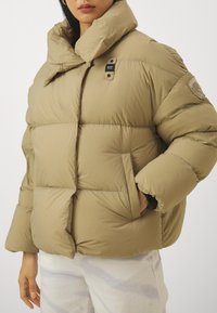 Beige vadderad jacka med hög krage, stora vadderade sektioner, sidofickor och ett märke på ärmen. Mjuk textur och avslappnad passform.