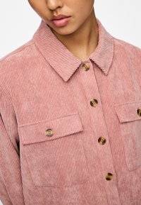 Jongere in een stoffige roze corduroy shirt met knoopzakken op de borst en schildpadmotief knopen, cropped bij de lippen en schouders.