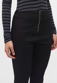 Personne portant un pantalon noir taille haute avec une fermeture éclair à l'avant et des poches, associé à un chemisier rayé noir et gris à manches longues.