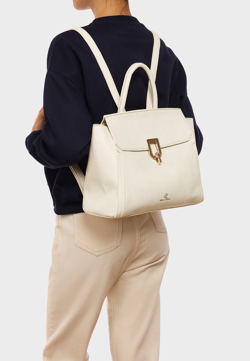 Zaino in pelle bianca texture con patta quadrata, hardware dorato e doppi manici superiori. Indossato sulla schiena sopra un maglione scuro.