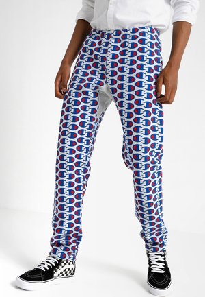 Personne portant une chemise blanche, un pantalon blanc et bleu avec un motif répétitif du logo Champion, et des baskets montantes à carreaux noirs et blancs.