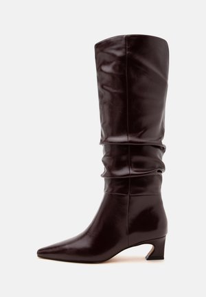 Botte en cuir marron foncé mi-mollet avec un bout pointu, une tige souple et un petit talon effilé sur un fond clair uni.