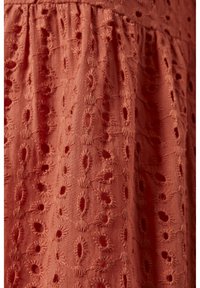 Látka v rzi barvě s texturovaným, vyšívaným vzorem obsahujícím oválné výřezy a kruhové akcenty, vytvářející jemný, podrobný design.