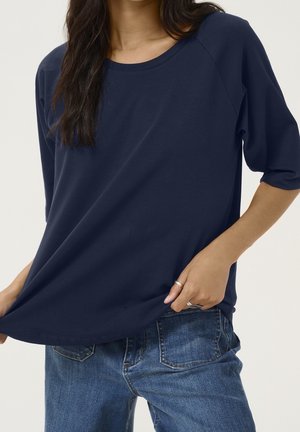 Femme portant un haut bleu marine à manches trois-quarts et un jean en denim bleu, tenant l'ourlet du haut d'une main.