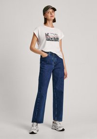 Camiseta gráfica blanca con un corte relajado, combinada con jeans de mezclilla rectos de tiro alto en azul oscuro. Detalles: zapatillas gris y gorra oliva.