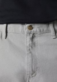 Pantalon gris avec un bouton en laiton fermé à la taille, porté avec une chemise noire rentrée.