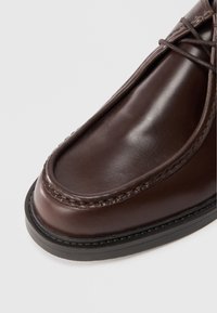 Brūna ādas kurpe ar gludu apdari, noapaļu pirkstu, kontrastējošiem vīlējumiem un cietu melnu gumijas zoli.