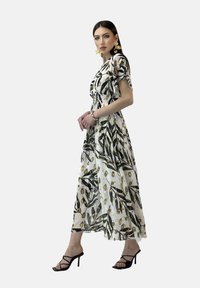 Robe maxi florale blanche avec des motifs verts et jaunes. Manches courtes et corsage ajusté, jupe fluide, complétée par des sandales noires.