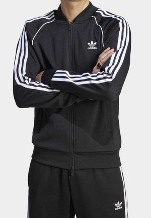 Personne portant une veste et un pantalon de survêtement Adidas noirs avec des bandes blanches et des logos, les bras croisés sur la poitrine.