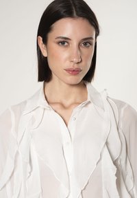 Blusa bianca e trasparente con un design a ruches, chiusura sul davanti con bottoni e colletto. La texture morbida e gli accenti a strati migliorano il suo aspetto.