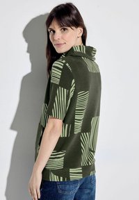 Maglietta verde oliva a maniche corte con scollo cowl, caratterizzata da strisce testurizzate e motivi geometrici in verde chiaro. Indossata con jeans in denim.