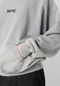 Sudadera gris con un ajuste relajado, que presenta un bolsillo frontal y un logo negro. El material parece suave con una textura ligeramente desgastada.