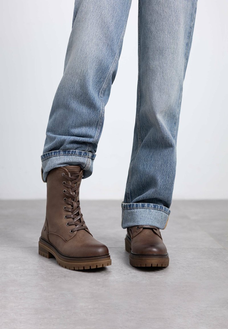 Botas marrones con cordones hechas de cuero, con una suela de goma gruesa y un acabado texturizado. Llevadas con unos jeans denim de color azul claro arremangados.