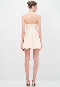 Robe crème sans bretelles avec un corsage ajusté, une jupe évasée et une fermeture éclair à l'arrière. Portée avec des talons à lanières, mettant en valeur une texture lisse et des lignes épurées.