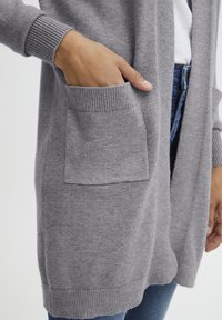 Šedý cardigan vyrobený z měkké pleteniny, s manžetami na žebro a velkou přední kapsou. Ležérní design s uvolněným střihem.