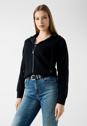 Jeune femme portant un sweat à capuche noir zippé avec cordons et un jean bleu avec une ceinture noire, debout devant un fond uni.