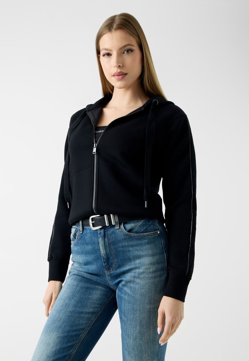 Jeune femme portant un sweat à capuche noir zippé avec cordons et un jean bleu avec une ceinture noire, debout devant un fond uni.