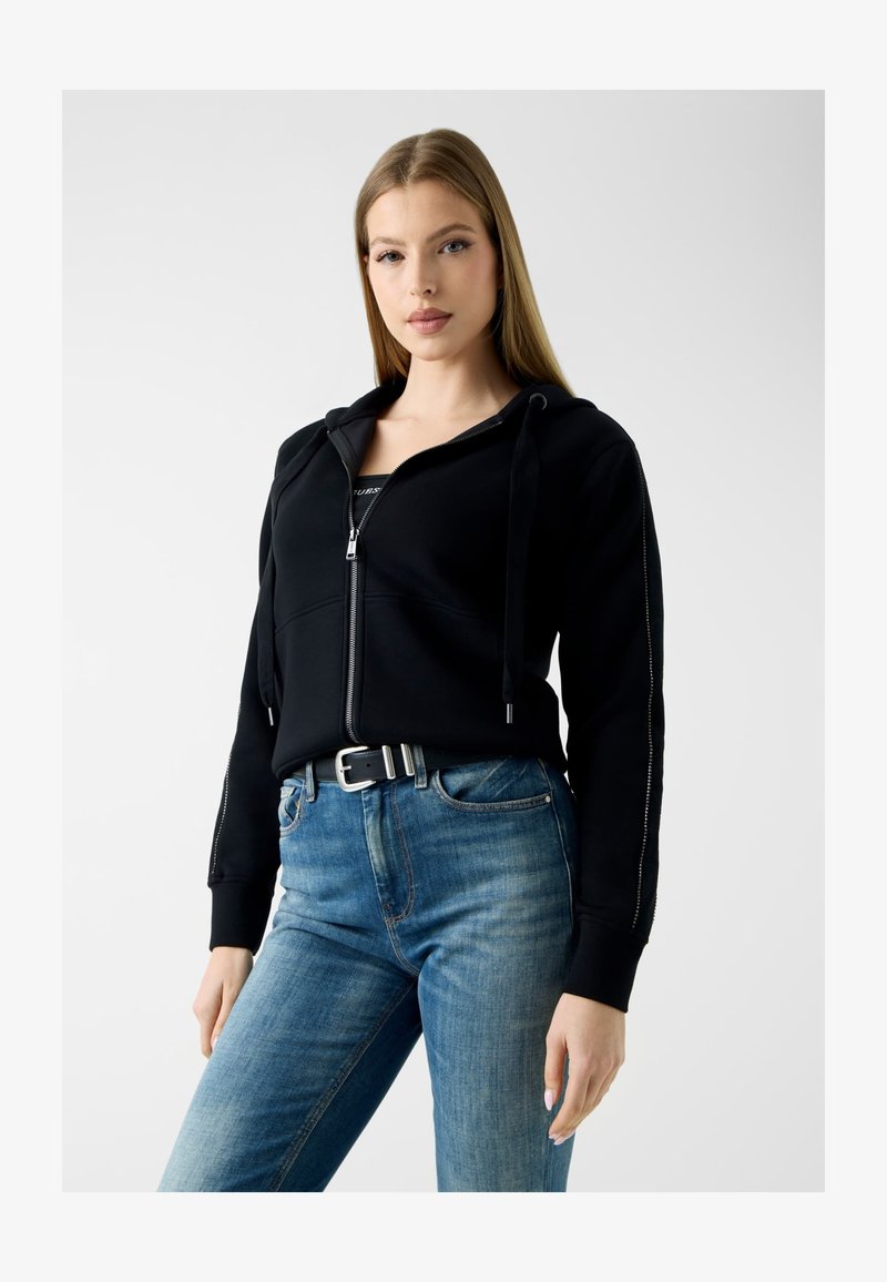 Jeune femme portant un sweat à capuche noir zippé avec cordons et un jean bleu avec une ceinture noire, debout devant un fond uni.