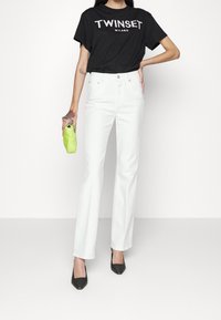 Chemise noire en coton avec le texte "TWINSET MILANO", associée à un jean flare blanc taille haute, et tenant une pochette verte citron.