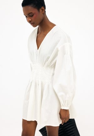 Žena s krátkymi vlasmi v bielych dlhých rukávoch a šatách so zúženým pásom, držiaca čiernu textúrovanú kabelku na jednoduchom pozadí.