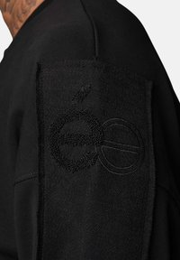 Sweatshirt noir avec un tissu texturé sur les épaules, comportant des détails de logo brodés, des coutures contrastées et une coupe près du corps.