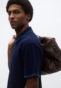 Μπλε πουκάμισο με υφή και μπλε ραφές στο γιακά, φορεμένο από άτομο που κουβαλά καφέ δερμάτινη τσάντα με κυκλικό μεταλλικό στοιχείο.