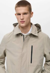 Giacca formale beige con cappuccio, chiusura frontale con zip e una tasca laterale con zip. Tessuto liscio, design minimalista, tessuto leggero.